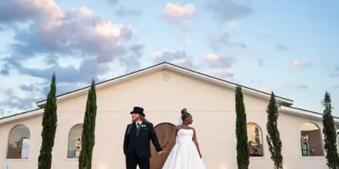 A bride and groom holding hands in front of a white building with tall cypress trees under a cloudy sky.