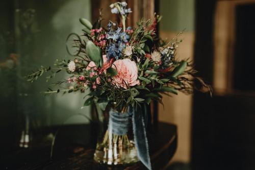 A close-up photograph of a mixed floral arrangement with pink and blue flowers and greenery in a glass vase.