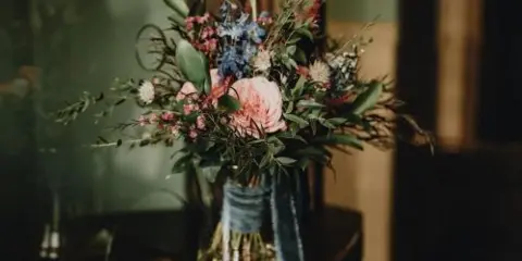A close-up photograph of a mixed floral arrangement with pink and blue flowers and greenery in a glass vase.