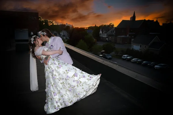 A bride and groom in wedding attire share a kiss during a dip pose on a rooftop at sunset, with a town and church steeple in the background.