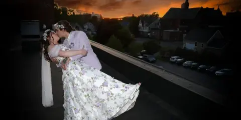 A bride and groom in wedding attire share a kiss during a dip pose on a rooftop at sunset, with a town and church steeple in the background.