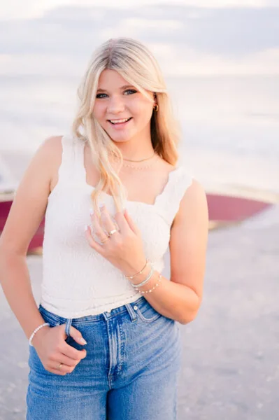 A professional portrait of a young woman with blonde hair, wearing a white top and jeans, standing outdoors near a body of water.
