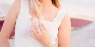 A professional portrait of a young woman with blonde hair, wearing a white top and jeans, standing outdoors near a body of water.
