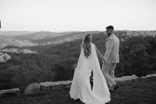 A black and white photograph of a bride and groom holding hands and looking at each other on a scenic hilltop overlooking a valley.