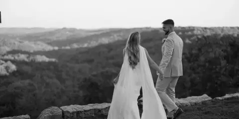 A black and white photograph of a bride and groom holding hands and looking at each other on a scenic hilltop overlooking a valley.