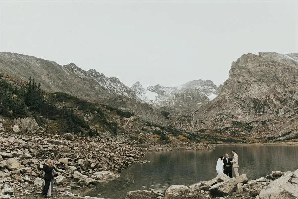 A bride and groom standing by a mountain lake during a wedding ceremony with an officiant.