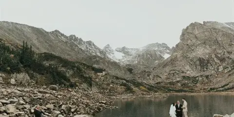 A bride and groom standing by a mountain lake during a wedding ceremony with an officiant.