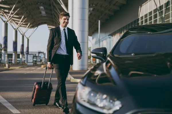 A man in a black suit and tie pulls a rolling suitcase next to a black luxury sedan at an airport curb.