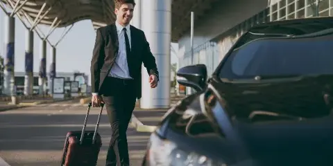 A man in a black suit and tie pulls a rolling suitcase next to a black luxury sedan at an airport curb.
