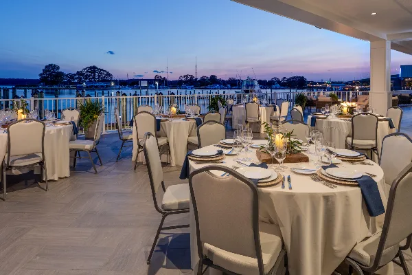 Formal dining tables set on a waterfront patio overlooking a marina at dusk.