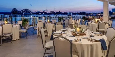 Formal dining tables set on a waterfront patio overlooking a marina at dusk.