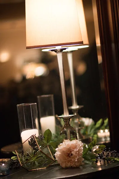 A close-up of an elegantly styled event table featuring a tall lamp, pillar candles in glass holders, a geometric terrarium, and a pink peony flower.