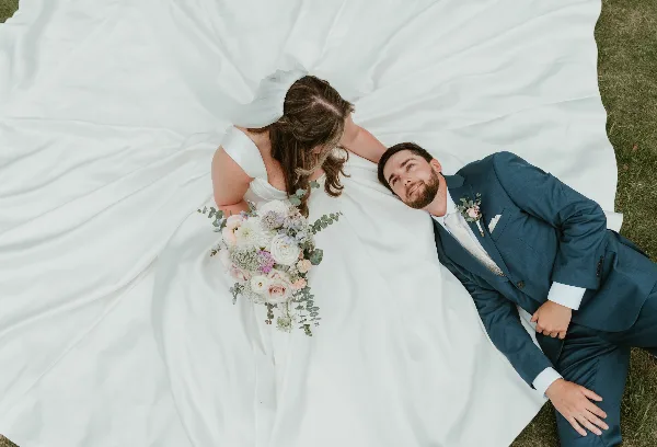 Overhead view of a bride and groom lying on grass, the bride in a white gown holding a bouquet and the groom in a blue suit.