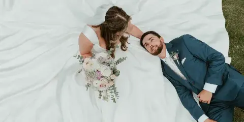 Overhead view of a bride and groom lying on grass, the bride in a white gown holding a bouquet and the groom in a blue suit.