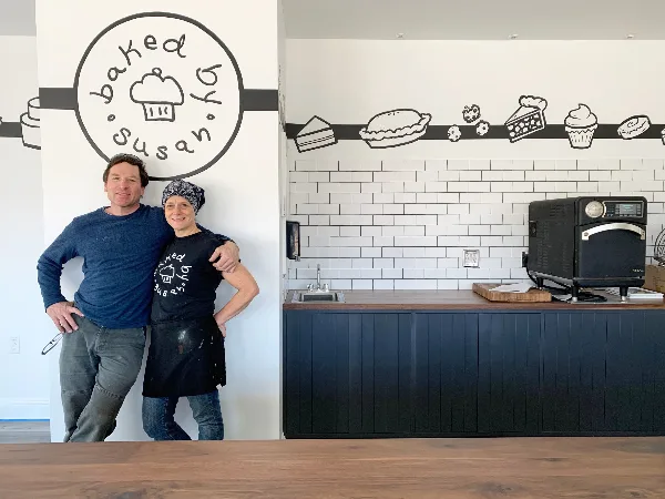 Two people standing in a bakery interior with a 'Baked by Susan' logo on the wall and a commercial oven on a counter.