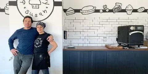 Two people standing in a bakery interior with a 'Baked by Susan' logo on the wall and a commercial oven on a counter.