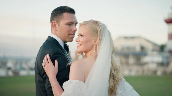 A bride and groom standing close together outdoors, with the groom in a black suit and the bride in a white lace dress and veil.