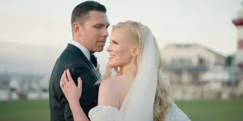 A bride and groom standing close together outdoors, with the groom in a black suit and the bride in a white lace dress and veil.