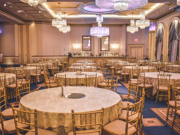 A spacious banquet hall set up for an event with round tables, gold chairs, and crystal chandeliers.