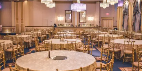 A spacious banquet hall set up for an event with round tables, gold chairs, and crystal chandeliers.