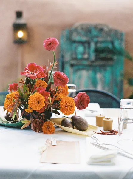 A vibrant floral arrangement featuring orange marigolds and red ranunculus on a white tablecloth, accompanied by candles and tableware.