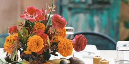 A vibrant floral arrangement featuring orange marigolds and red ranunculus on a white tablecloth, accompanied by candles and tableware.