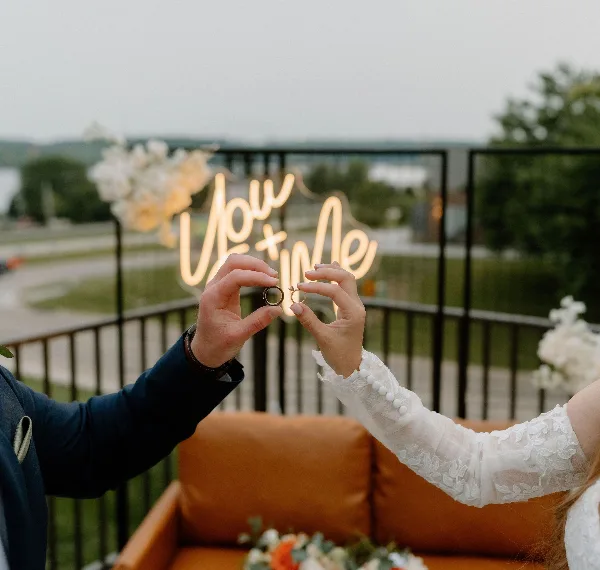 A couple holding wedding rings in front of a glowing neon 'You + Me' sign, with white floral arrangements and a tan sofa in the background.