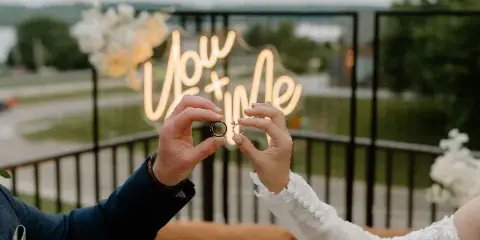 A couple holding wedding rings in front of a glowing neon 'You + Me' sign, with white floral arrangements and a tan sofa in the background.