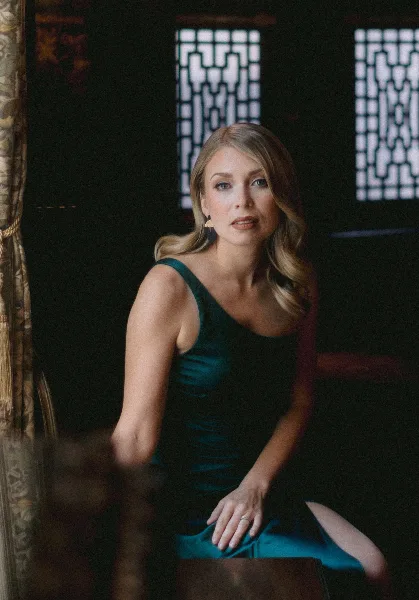 A woman in a teal dress sitting in a dimly lit room with traditional window patterns in the background.