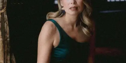 A woman in a teal dress sitting in a dimly lit room with traditional window patterns in the background.