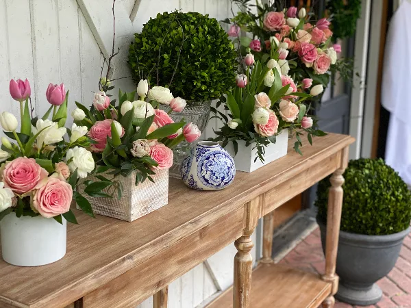 A wooden console table displaying multiple floral arrangements with pink and white roses, tulips, and boxwood topiaries.