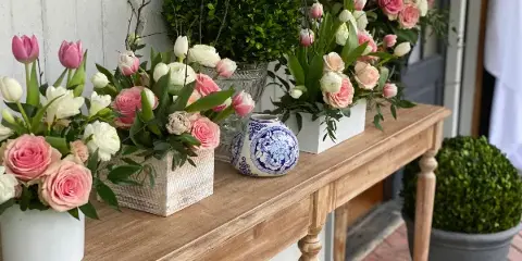 A wooden console table displaying multiple floral arrangements with pink and white roses, tulips, and boxwood topiaries.