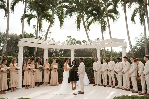 A wedding ceremony taking place outdoors under a white pergola structure with palm trees in the background.