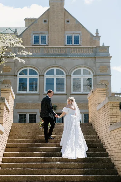 A bride and groom holding hands on the stone steps of a large brick building.