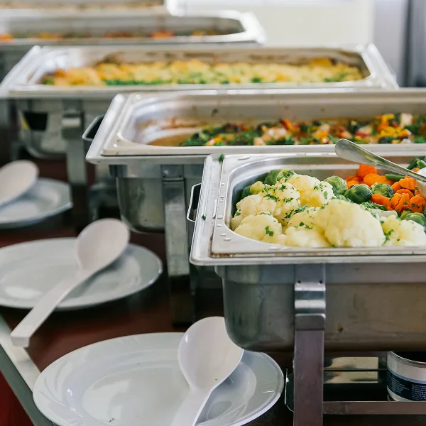 A catering buffet line featuring stainless steel chafing dishes filled with prepared food, including cauliflower, Brussels sprouts, and carrots, with serving spoons and plates ready for guests.