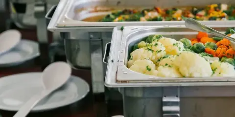 A catering buffet line featuring stainless steel chafing dishes filled with prepared food, including cauliflower, Brussels sprouts, and carrots, with serving spoons and plates ready for guests.