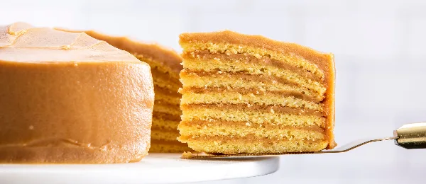 A close-up photograph of a multi-layered caramel cake with a slice being lifted by a server, showing the distinct layers of sponge and caramel icing.