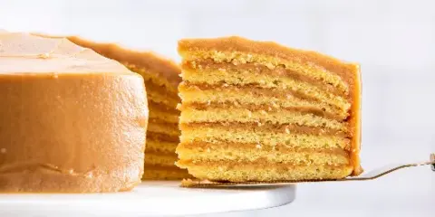 A close-up photograph of a multi-layered caramel cake with a slice being lifted by a server, showing the distinct layers of sponge and caramel icing.