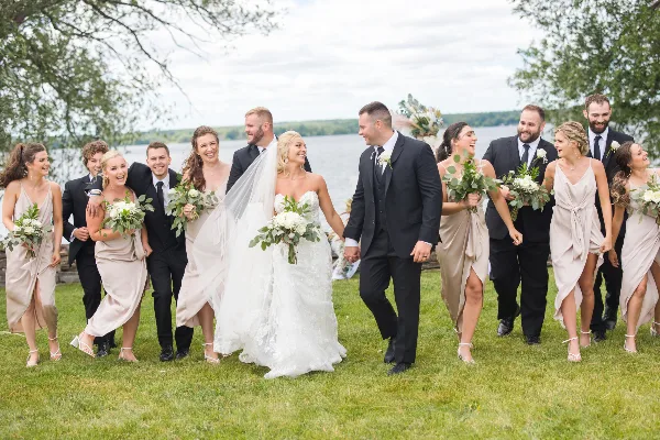 A wedding party consisting of a bride, groom, and attendants walking across a grassy lawn with a river visible in the background.