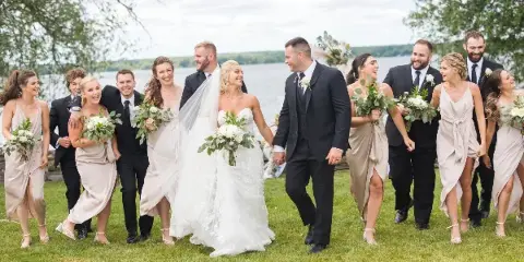 A wedding party consisting of a bride, groom, and attendants walking across a grassy lawn with a river visible in the background.