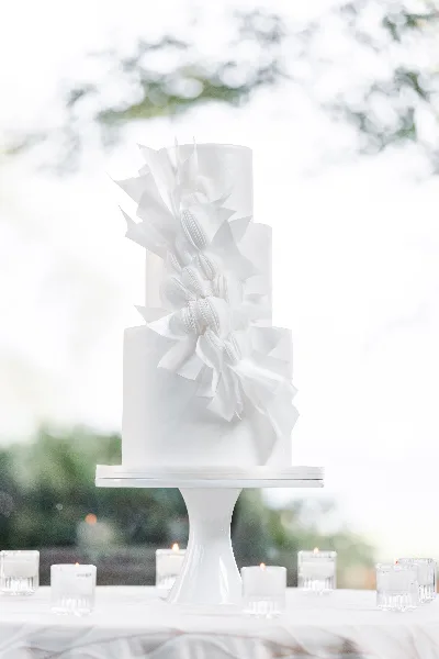 A tall, white, three-tiered wedding cake decorated with cascading white sugar flowers, displayed on a white pedestal stand.