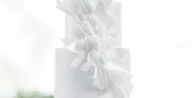 A tall, white, three-tiered wedding cake decorated with cascading white sugar flowers, displayed on a white pedestal stand.