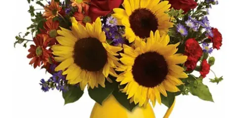 A floral arrangement featuring sunflowers, red roses, and purple flowers in a yellow pitcher vase.