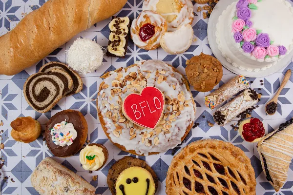 An overhead shot of an assortment of baked goods including a baguette, cinnamon rolls, a decorated cake, a fruit pie, and various pastries arranged on a patterned surface.