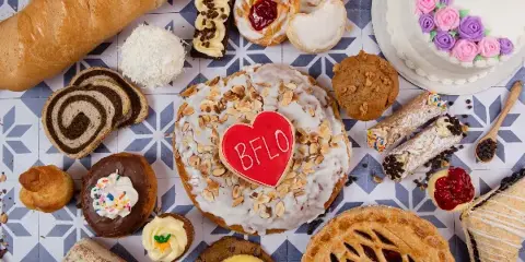 An overhead shot of an assortment of baked goods including a baguette, cinnamon rolls, a decorated cake, a fruit pie, and various pastries arranged on a patterned surface.