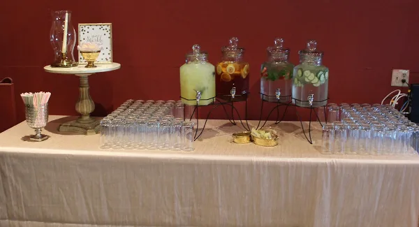 A catering beverage station featuring glass dispensers with infused drinks and rows of empty glasses on a table.