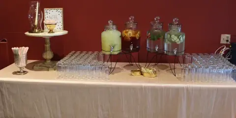A catering beverage station featuring glass dispensers with infused drinks and rows of empty glasses on a table.