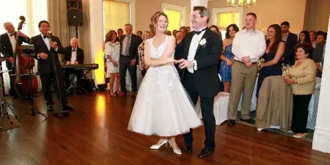 A bride and groom dancing at a wedding reception with guests and a live band in the background.