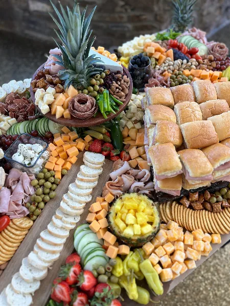 A large catering spread featuring a charcuterie board with meats and cheeses, a row of sandwiches, and fresh fruit.