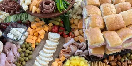 A large catering spread featuring a charcuterie board with meats and cheeses, a row of sandwiches, and fresh fruit.